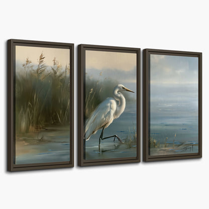 A white heron beside tall marsh grasses and still blue water 3 piece framed canvas wall art on a white background