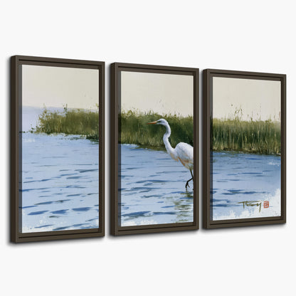 A white heron wading through bright blue water beside green reeds 3 piece framed canvas wall art on a white background