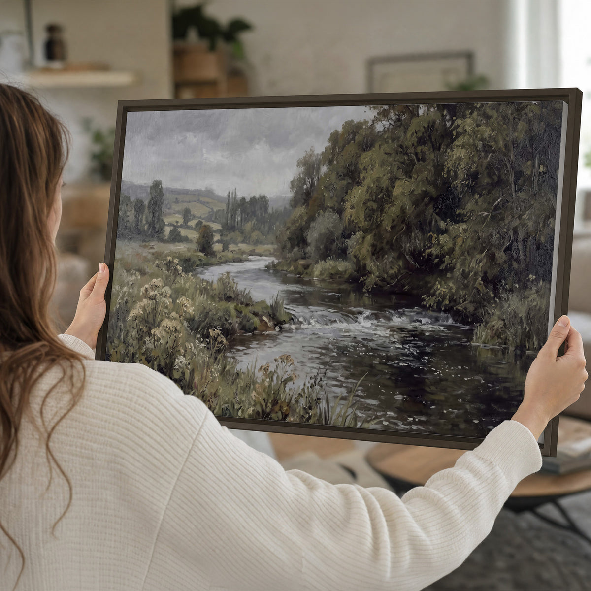 A gently flowing river bordered by olive-green trees and flowers single framed canvas wall art held in hands