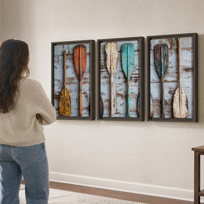 A row of colorful weathered wooden oars against pale blue boards 3 piece framed canvas wall art three panel arrangement on a wall