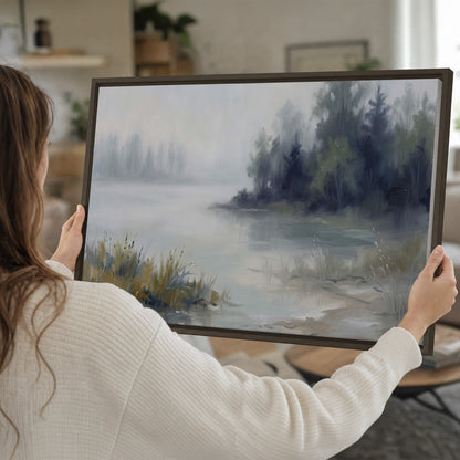 A calm pale lake bordered by dark evergreen trees and distant mist single framed canvas wall art held in hands