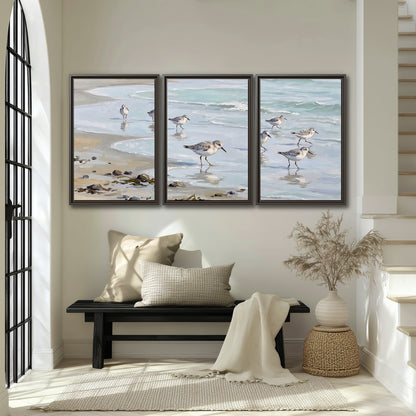 Small shorebirds standing along a pale blue shoreline 3 piece framed canvas wall art displayed in a entryway