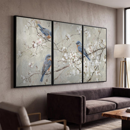 Bluebirds perched on delicate white flowering branches against pale sage 3 piece framed canvas wall art displayed in a modern living room
