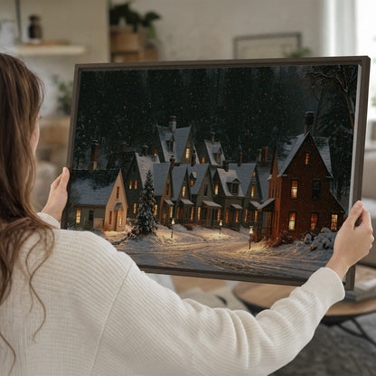 Snow-covered village cottages with glowing windows beneath a dark evergreen forest single framed canvas wall art held in hands