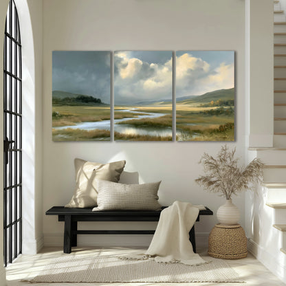 A winding stream through an open meadow beneath large sunlit clouds 3 piece canvas wall art displayed in a entryway