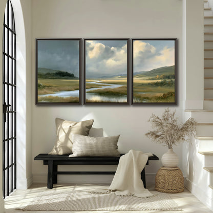 A winding stream through an open meadow beneath large sunlit clouds 3 piece framed canvas wall art displayed in a entryway