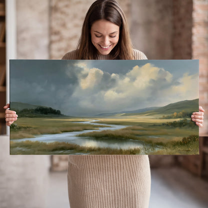 A winding stream through an open meadow beneath large sunlit clouds single canvas wall art held in hands