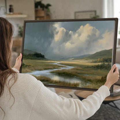 A winding stream through an open meadow beneath large sunlit clouds single framed canvas wall art held in hands