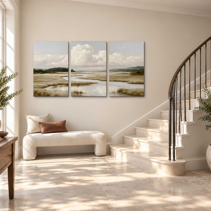 A pale winding waterway crossing quiet wetlands beneath billowing white clouds 3 piece canvas wall art styled in an interior