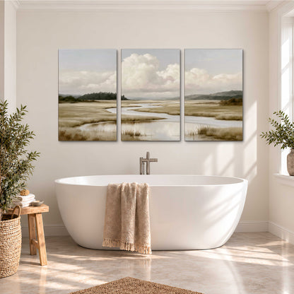 A pale winding waterway crossing quiet wetlands beneath billowing white clouds 3 piece canvas wall art styled in an interior