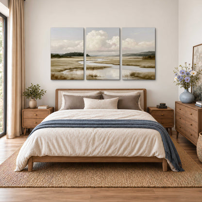 A pale winding waterway crossing quiet wetlands beneath billowing white clouds 3 piece canvas wall art styled in an interior