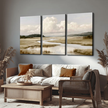 A pale winding waterway crossing quiet wetlands beneath billowing white clouds 3 piece canvas wall art displayed in a rustic living room