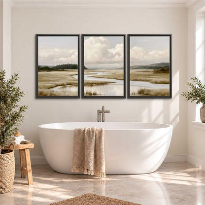 A pale winding waterway crossing quiet wetlands beneath billowing white clouds 3 piece framed canvas wall art styled in an interior