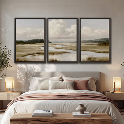 A pale winding waterway crossing quiet wetlands beneath billowing white clouds 3 piece framed canvas wall art displayed in a bedroom