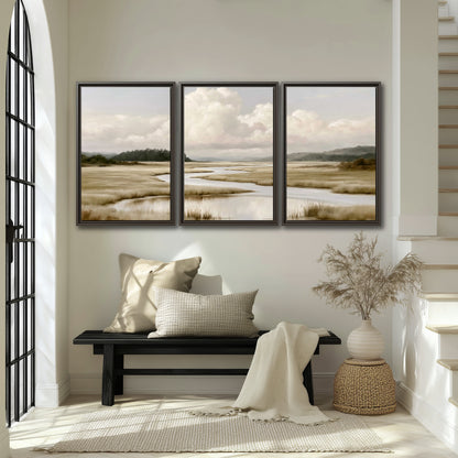 A pale winding waterway crossing quiet wetlands beneath billowing white clouds 3 piece framed canvas wall art displayed in a entryway