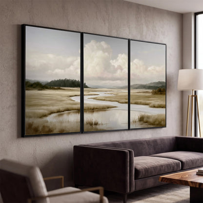 A pale winding waterway crossing quiet wetlands beneath billowing white clouds 3 piece framed canvas wall art displayed in a modern living room