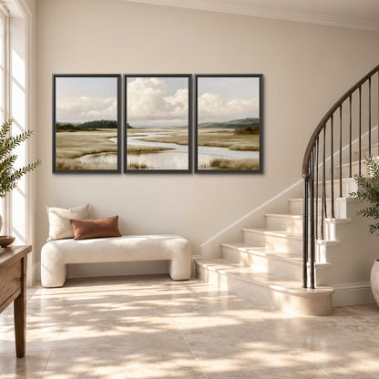 A pale winding waterway crossing quiet wetlands beneath billowing white clouds 3 piece framed canvas wall art styled in an interior