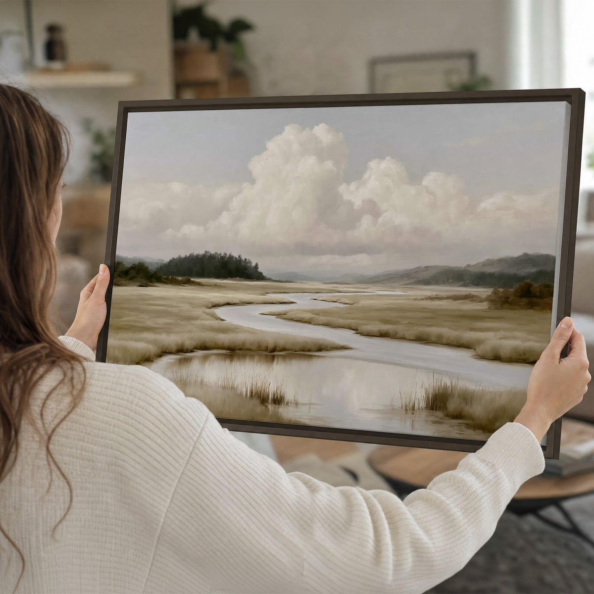 A pale winding waterway crossing quiet wetlands beneath billowing white clouds single framed canvas wall art held in hands