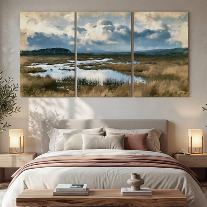 A wide open marsh with blue water and warm grasses beneath layered clouds 3 piece canvas wall art displayed in a bedroom