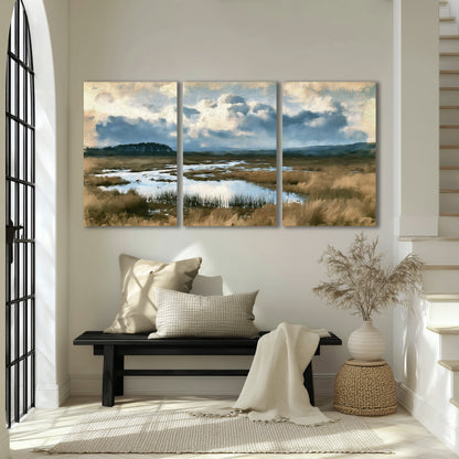 A wide open marsh with blue water and warm grasses beneath layered clouds 3 piece canvas wall art displayed in a entryway