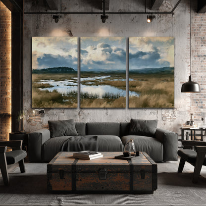 A wide open marsh with blue water and warm grasses beneath layered clouds 3 piece canvas wall art displayed in a industrial room