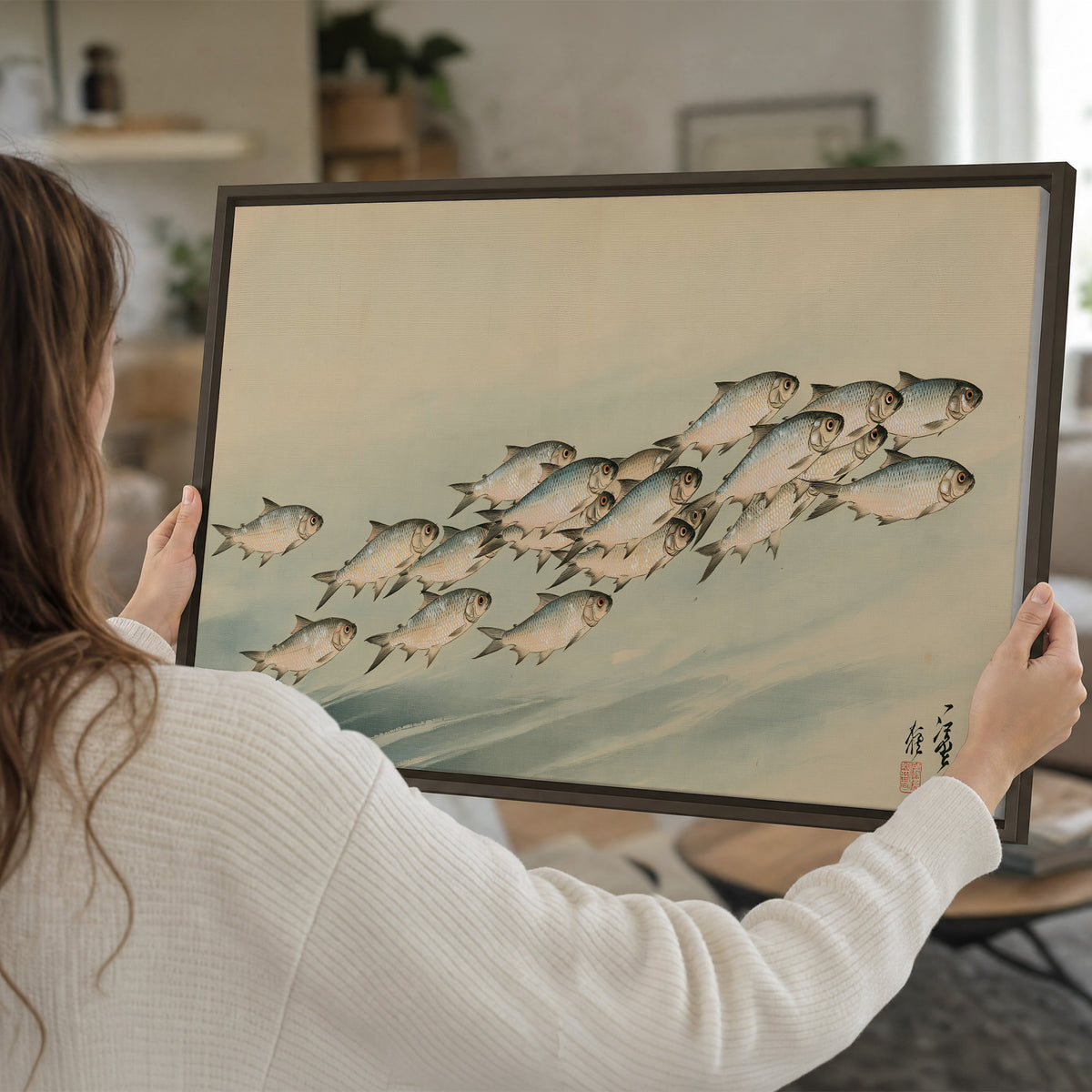 A flowing school of small fish above pale blue water on a cream ground single framed canvas wall art held in hands