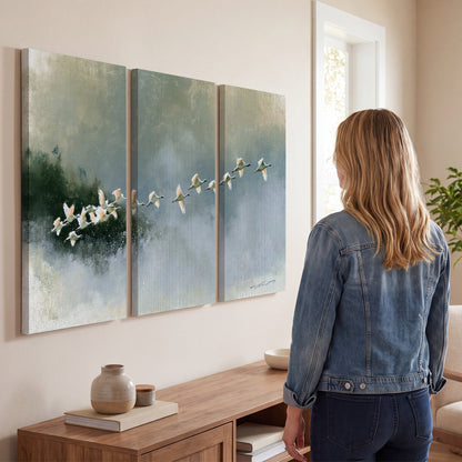 A flock of white birds flying low above a quiet blue-green lake 3 piece canvas wall art three panel arrangement on a wall