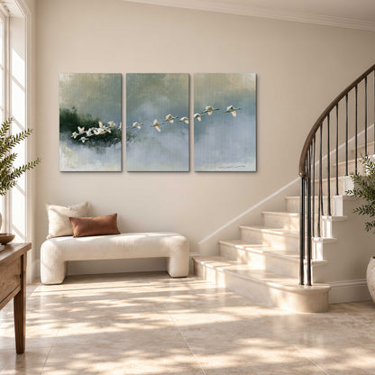 A flock of white birds flying low above a quiet blue-green lake 3 piece canvas wall art styled in an interior