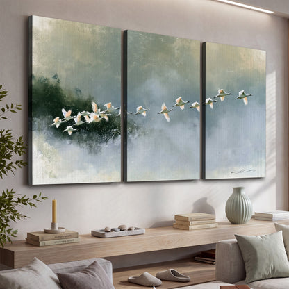 A flock of white birds flying low above a quiet blue-green lake 3 piece canvas wall art displayed in a console