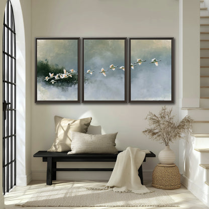 A flock of white birds flying low above a quiet blue-green lake 3 piece framed canvas wall art displayed in a entryway