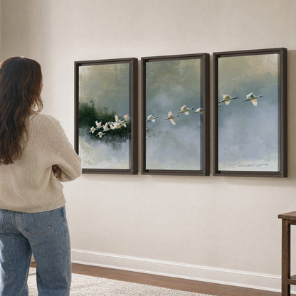 A flock of white birds flying low above a quiet blue-green lake 3 piece framed canvas wall art three panel arrangement on a wall