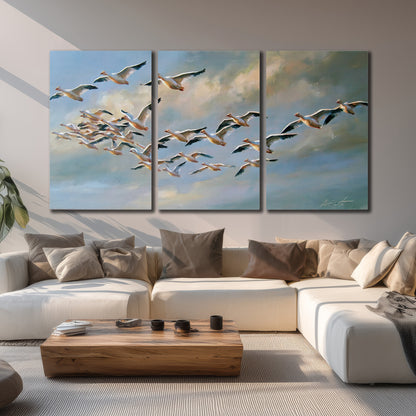 A flock of pale seabirds gliding beneath soft clouds across a blue sky 3 piece canvas wall art displayed in a living room