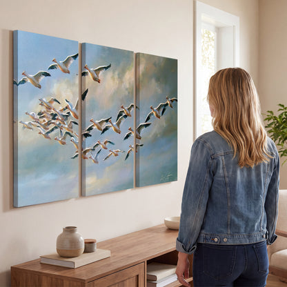 A flock of pale seabirds gliding beneath soft clouds across a blue sky 3 piece canvas wall art three panel arrangement on a wall