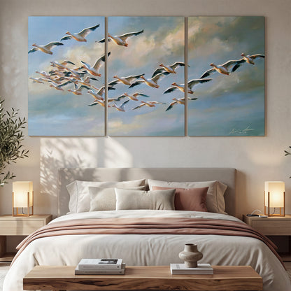 A flock of pale seabirds gliding beneath soft clouds across a blue sky 3 piece canvas wall art displayed in a bedroom