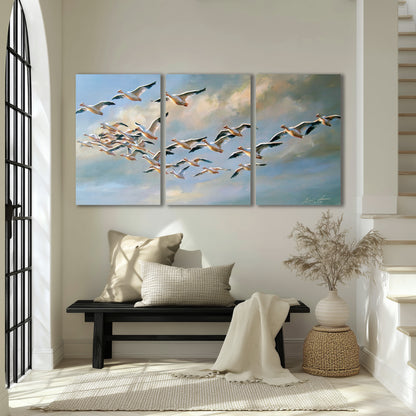 A flock of pale seabirds gliding beneath soft clouds across a blue sky 3 piece canvas wall art displayed in a entryway