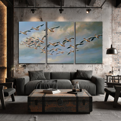 A flock of pale seabirds gliding beneath soft clouds across a blue sky 3 piece canvas wall art displayed in a industrial room