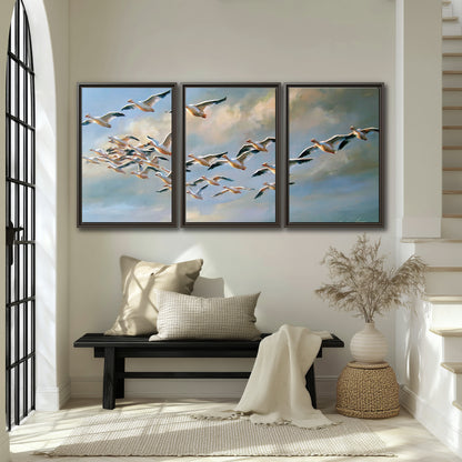 A flock of pale seabirds gliding beneath soft clouds across a blue sky 3 piece framed canvas wall art displayed in a entryway