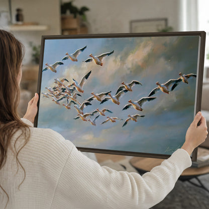 A flock of pale seabirds gliding beneath soft clouds across a blue sky single framed canvas wall art held in hands