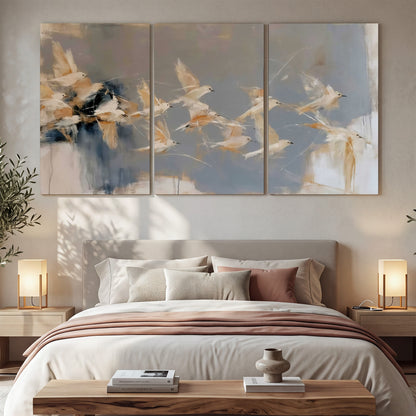Pale bird-like forms sweeping across layered strokes of blue, ochre and cream 3 piece canvas wall art displayed in a bedroom