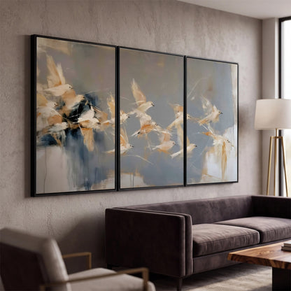 Pale bird-like forms sweeping across layered strokes of blue, ochre and cream 3 piece framed canvas wall art displayed in a modern living room