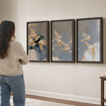 Pale bird-like forms sweeping across layered strokes of blue, ochre and cream 3 piece framed canvas wall art three panel arrangement on a wall