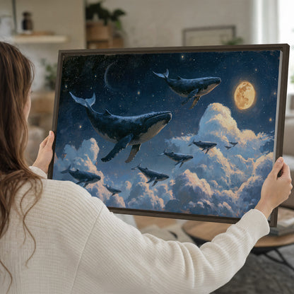 A group of whales floating among clouds beneath a golden moon in a deep blue night sky single framed canvas wall art held in hands