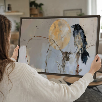 A dark raven beside a golden moon amid pale blue and cream painterly strokes single framed canvas wall art held in hands