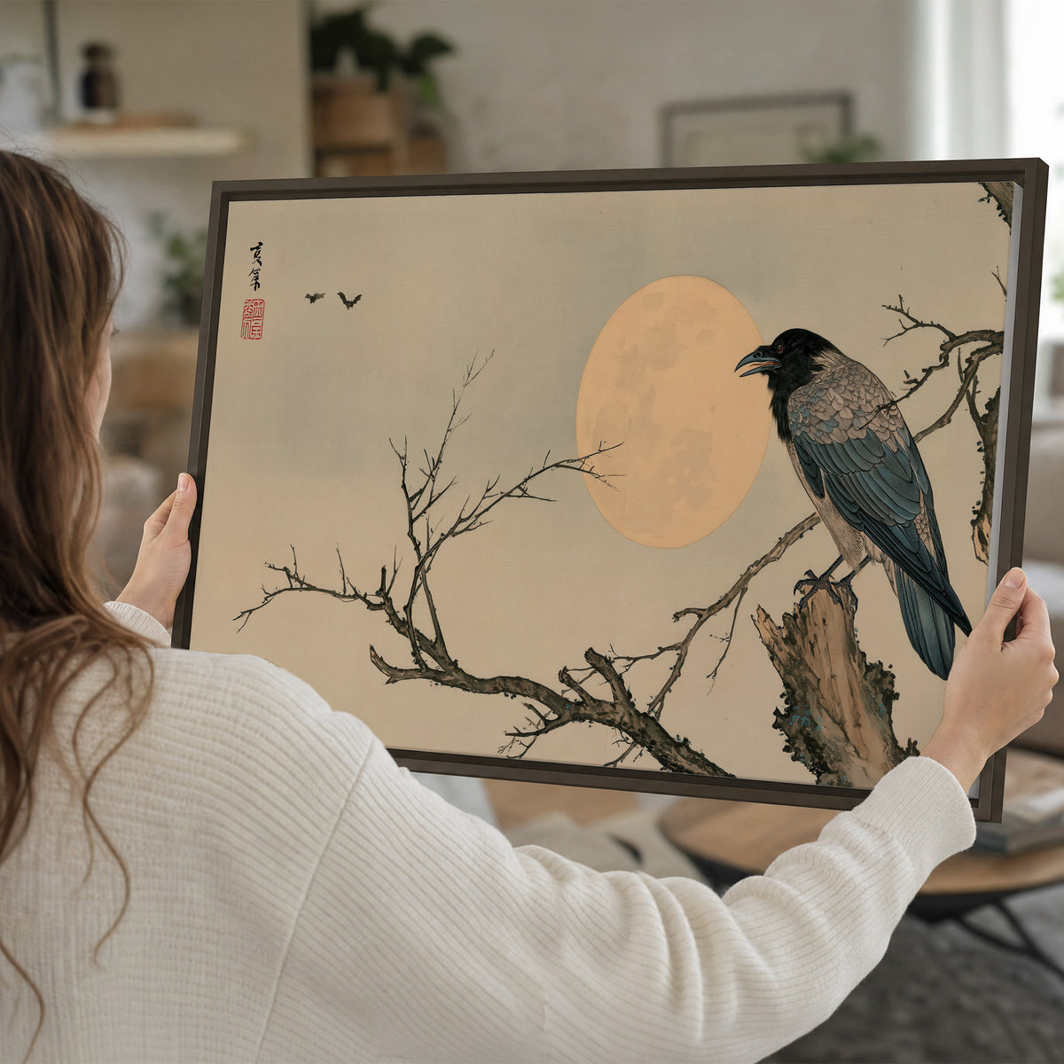 A dark raven perched on a bare branch beside a warm full moon single framed canvas wall art held in hands