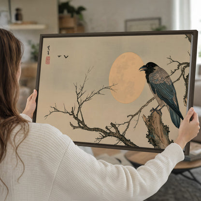 A dark raven perched on a bare branch beside a warm full moon single framed canvas wall art held in hands
