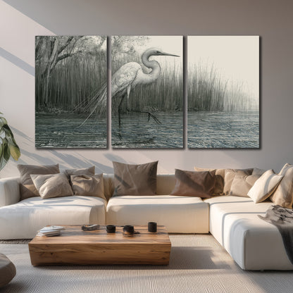 A pale long-legged heron standing beside tall reeds in a muted gray-green wetland 3 piece canvas wall art displayed in a living room