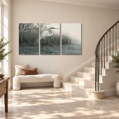 A pale long-legged heron standing beside tall reeds in a muted gray-green wetland 3 piece canvas wall art styled in an interior