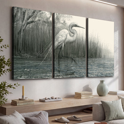 A pale long-legged heron standing beside tall reeds in a muted gray-green wetland 3 piece canvas wall art displayed in a console