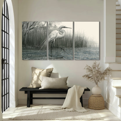 A pale long-legged heron standing beside tall reeds in a muted gray-green wetland 3 piece canvas wall art displayed in a entryway