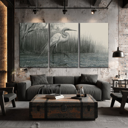 A pale long-legged heron standing beside tall reeds in a muted gray-green wetland 3 piece canvas wall art displayed in a industrial room